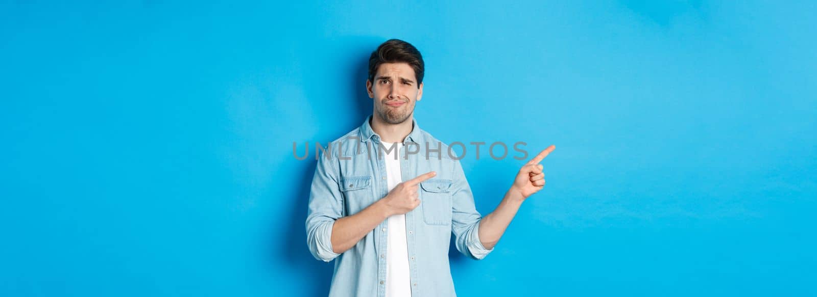 Skeptical adult man pointing fingers left, looking doubtful and unsure, standing over blue background by Benzoix
