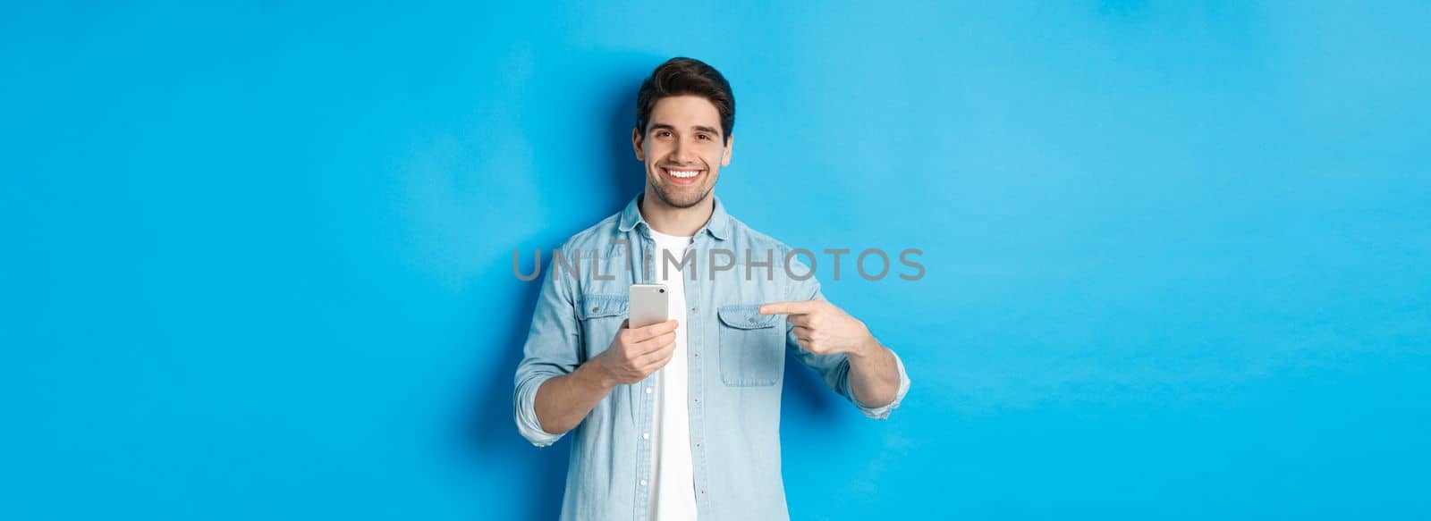 Concept of online shopping, applications and technology. Handsome man recommending app on smartphone, pointing at phone and smiling satisfied, standing over blue background.