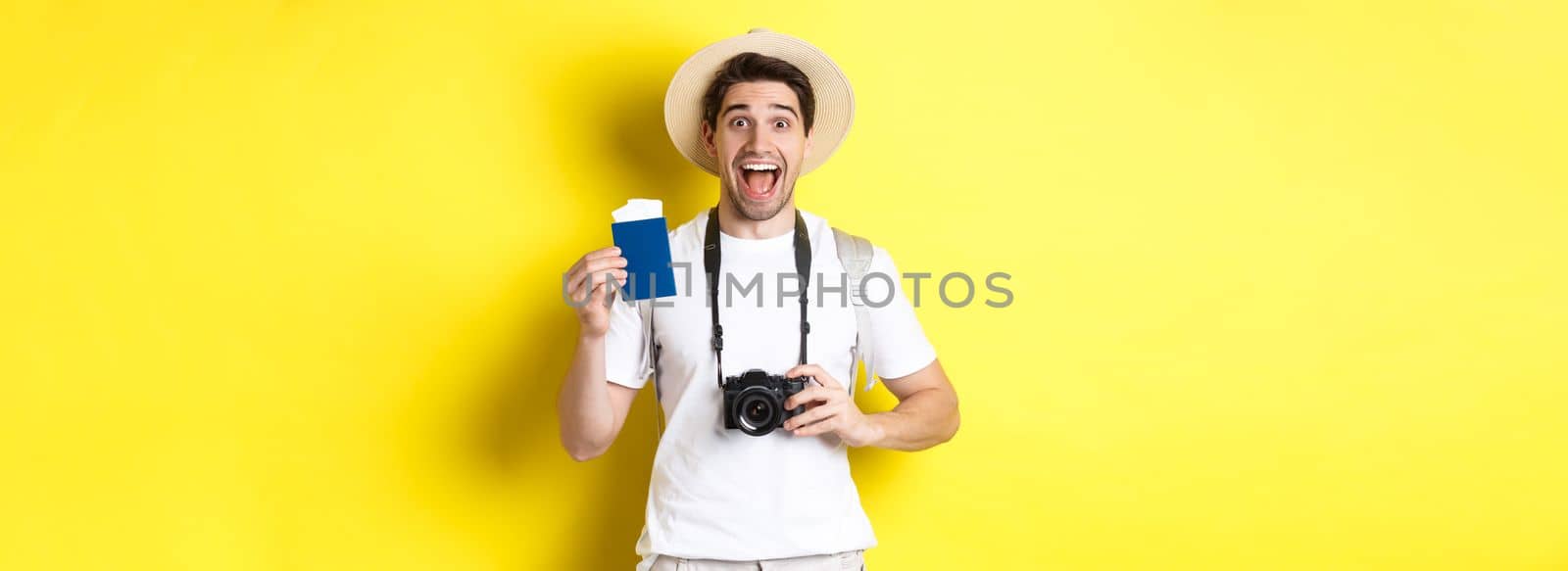 Travelling, vacation and tourism concept. Excoted tpirost showing passport with tickets, holding camera and wearing straw hat, standing over yellow background.
