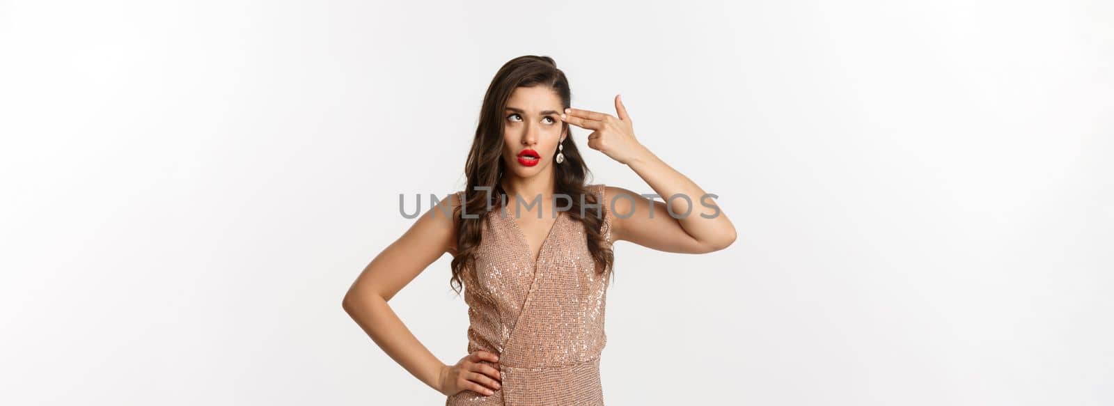 Celebration and party concept. Annoyed woman in elegant evening dress wants to kill herself from boredom, shooting finger gun in head and roll eyes irritated, standing over white background by Benzoix