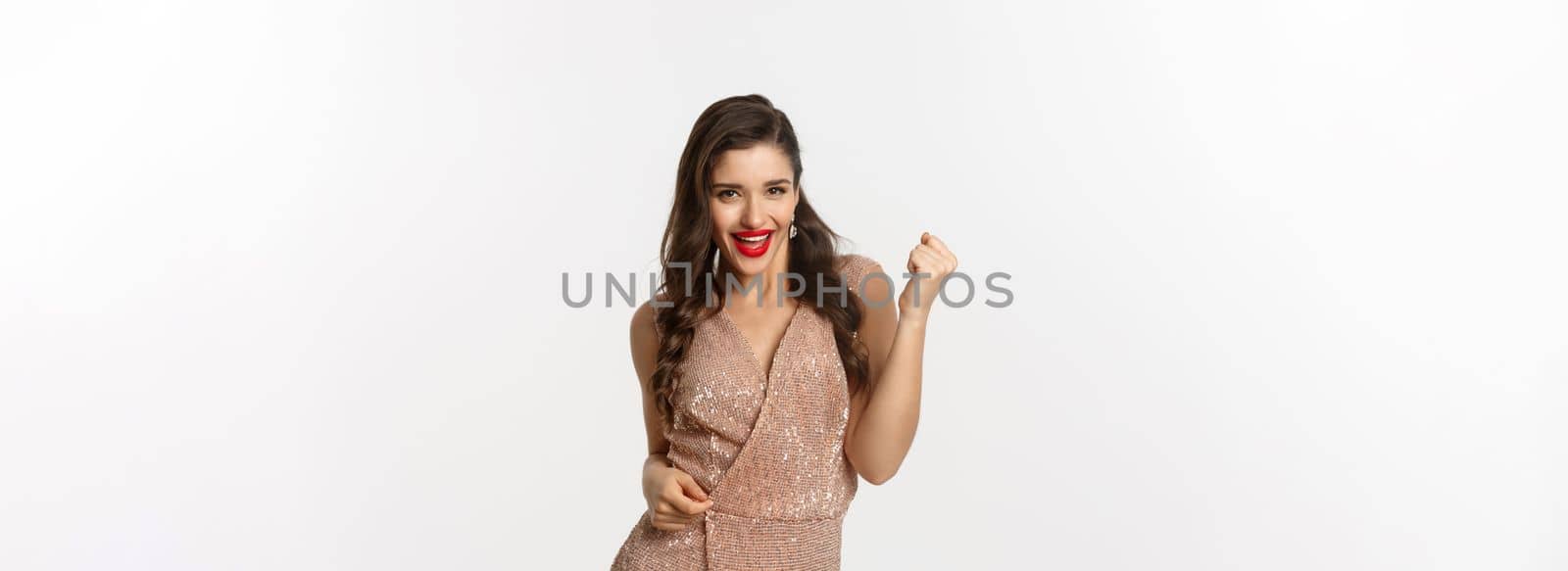 Celebration and party concept. Elegant young woman winning and feeling satisfied, wearing glamour dress, making fist pump and say yes, triumphing over white background.