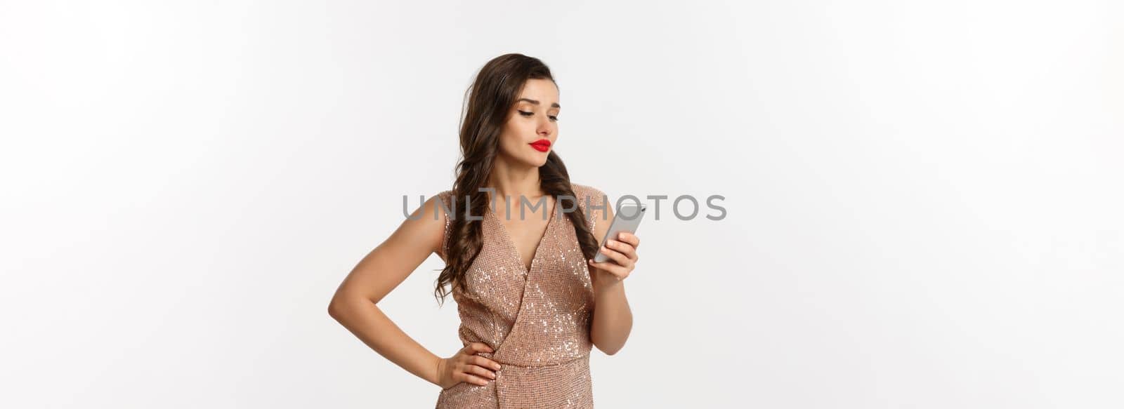 Christmas party and celebration concept. Careless woman in stylish dress reading message on mobile phone, looking skeptical, standing over white background.