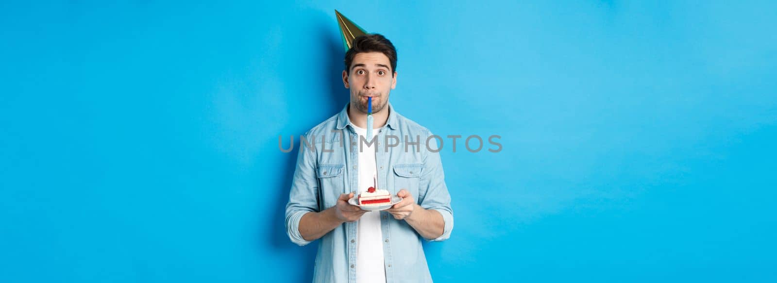 Handsome young man holding birthday cake, wearing party hat and blowing whistle, standing over blue background.
