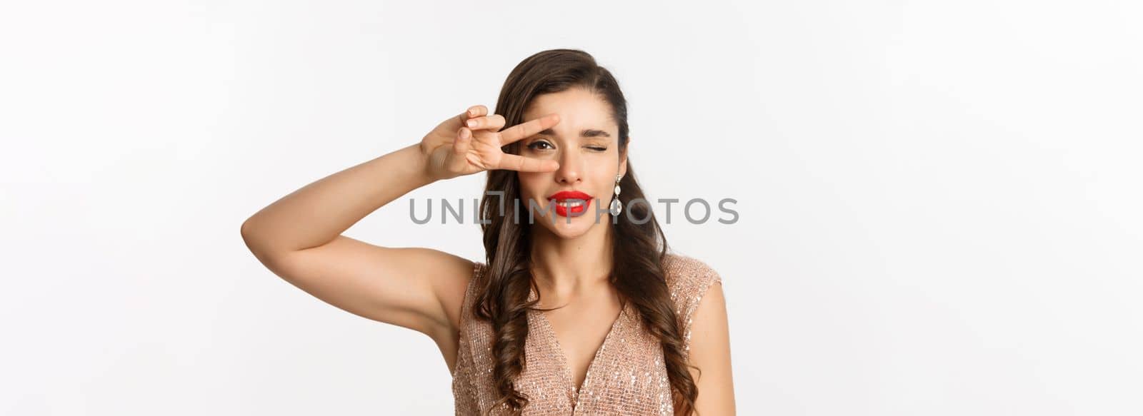 Concept of New Year celebration and winter holidays. Close-up of glamour woman in party dress, red lips, showing peace sign and winking sexy at camera, white background.