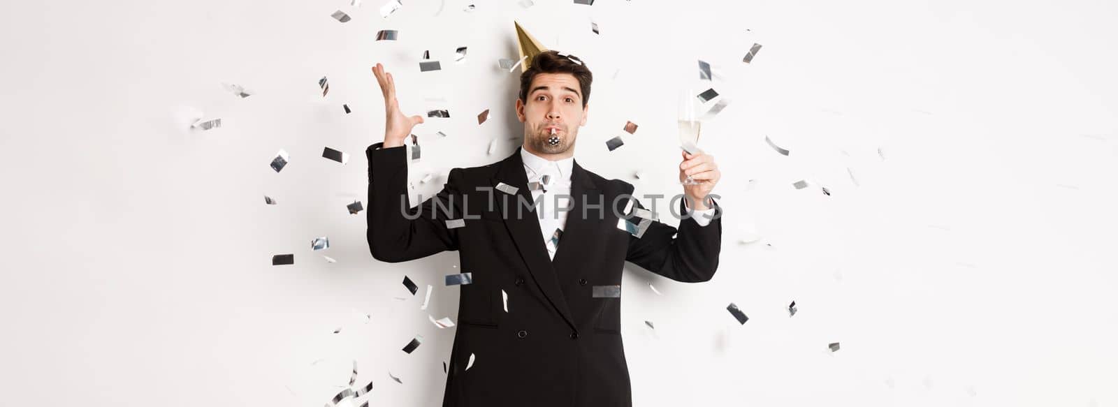 Handsome party guy in black suit having fun, celebrating new year, blowing whistle and drinking champagne while confetti falling, wishing happy holidays, standing against white background.