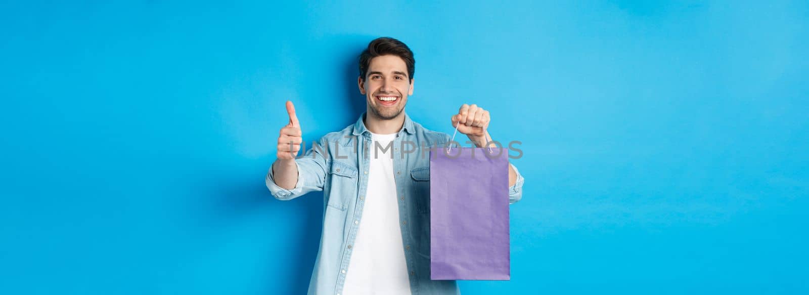 Concept of shopping, holidays and lifestyle. Satisfied smiling man holding paper bag, showing thumb-up and recommending store, standing over blue background.