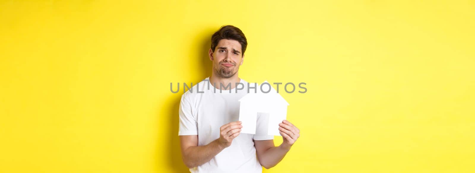 Real estate concept. Displeased young man showing paper house model and grimacing upset, standing over yellow background.