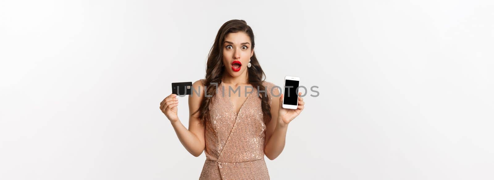 Online shopping and holidays concept. Woman looking amazed and showing credit card with smartphone screen, standing over white background.