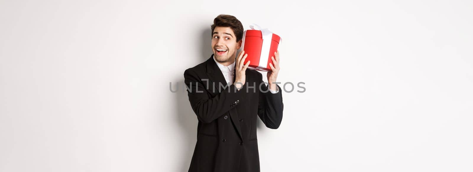 Concept of christmas holidays, celebration and lifestyle. Handsome man in black suit, shaking a box with gift, wondering whats inside, standing against white background.