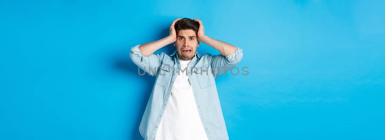 Image of man in panic holding hands on head, looking frustrated and anxious, standing against blue background.