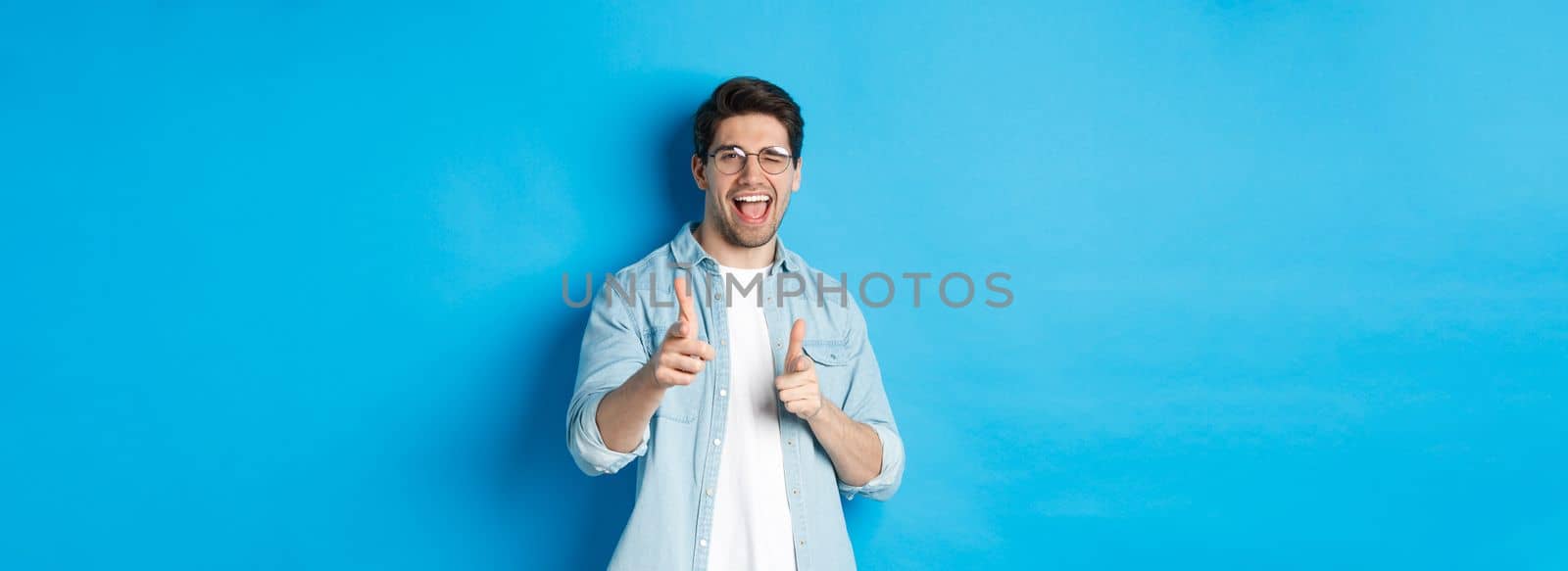 Confident man saying congrats, winking and pointing at you, standing pleased over blue background and smiling.