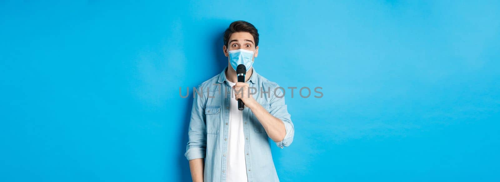 Young man in medical mask giving speech, holding microphone and looking confused, standing over blue background by Benzoix