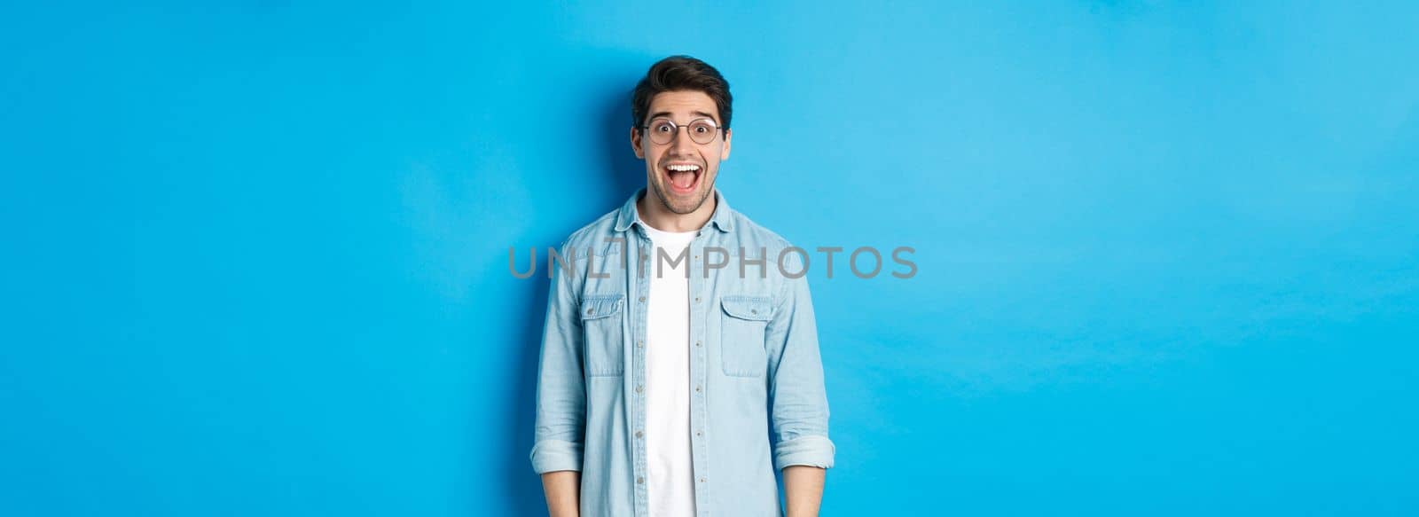 Attractive happy man in glasses looking surprised, checking out advertisement, standing over blue background by Benzoix