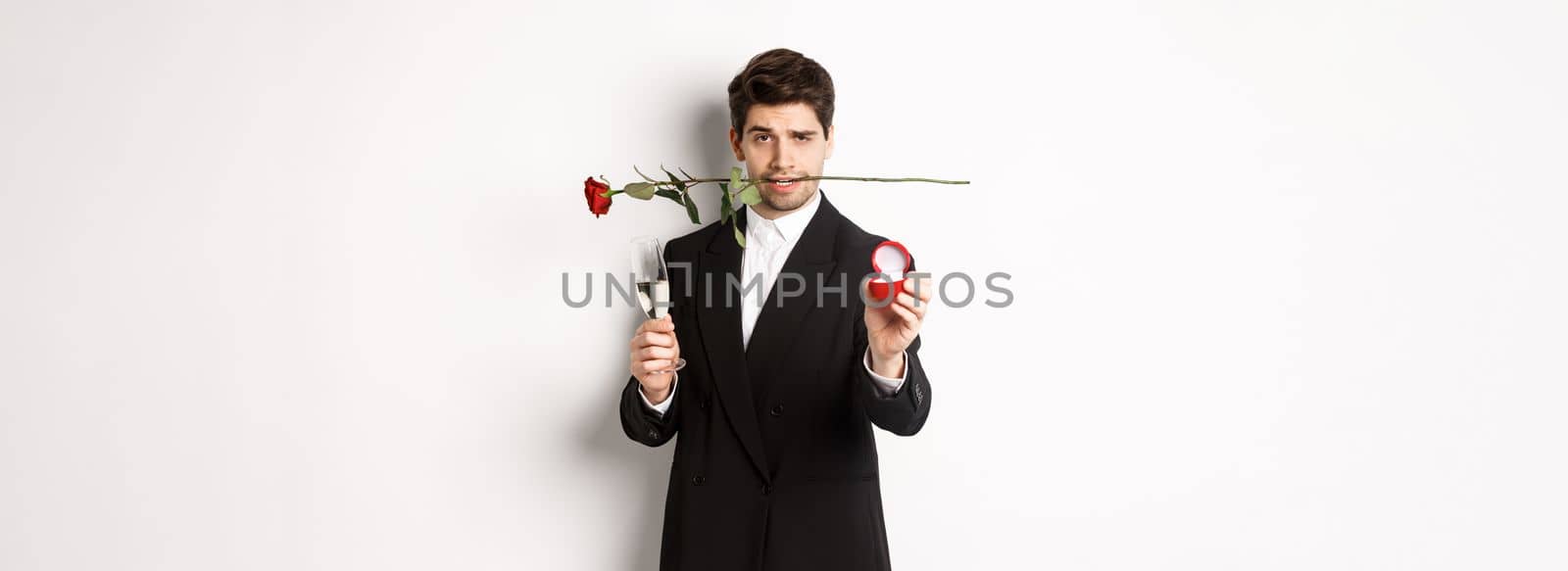 Passionate young man in suit making a proposal, holding rose in teeth and glass of champagne, showing engagement ring, asking to marry him, standing against white background by Benzoix