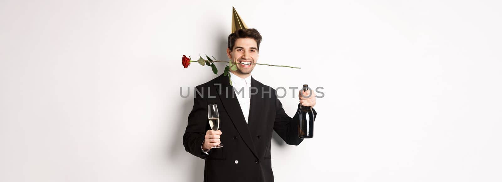 Funny guy in trendy suit, celebrating and having a party, holding rose in teeth and champagne, standing over white background.