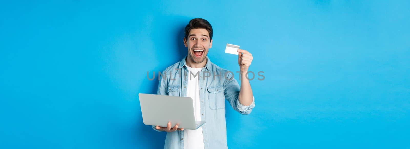 Excited man shop online, showing credit card and holding laptop, buying in internet, standing over blue background.
