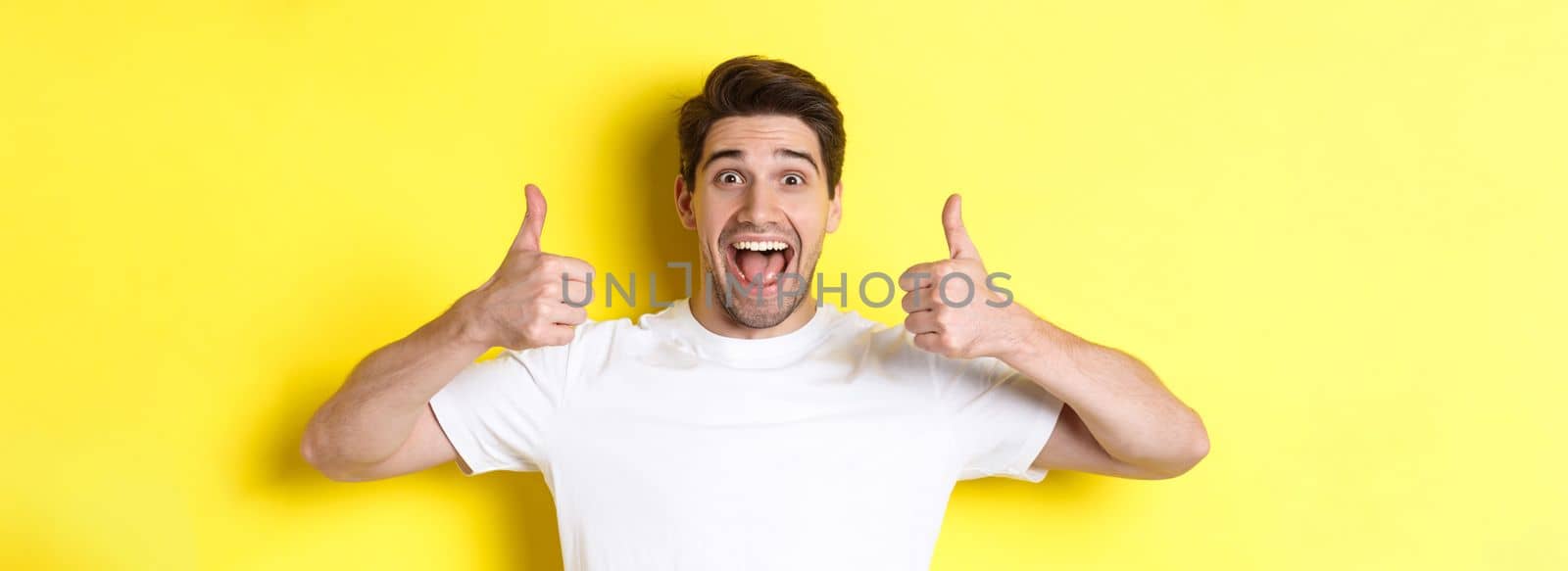 Close-up of handsome young man showing thumbs up, approve and agree, smiling satisfied, standing over yellow background.