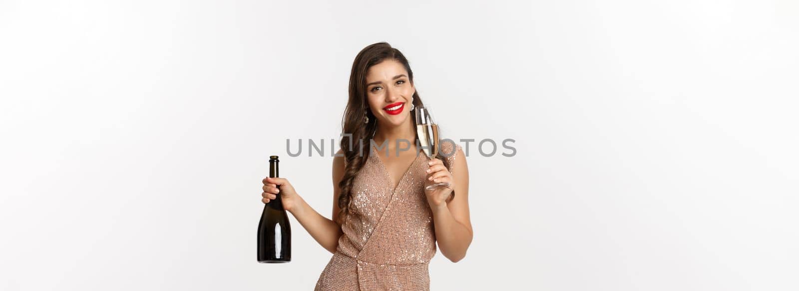 Winter holidays celebration concept. Image of happy young woman in luxury dress, drinking champagne from glass, holding bottle, New Year party, white background.