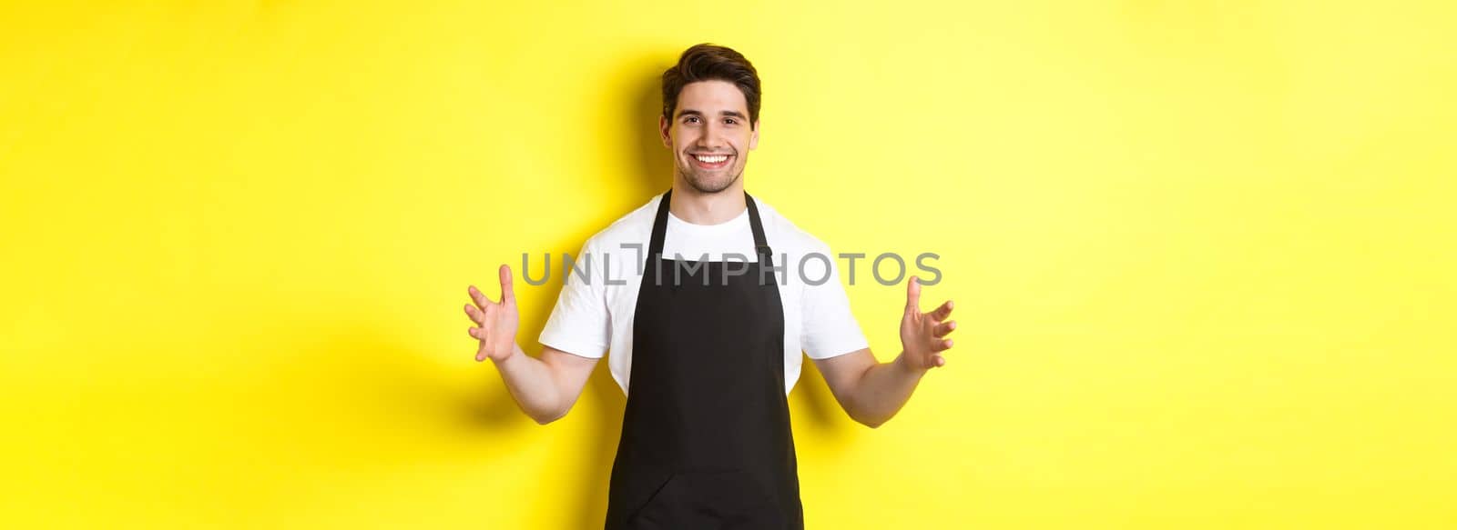 Smiling waiter in black apron holding your logo or box, spread hands as if carry something large, standing over yellow background.