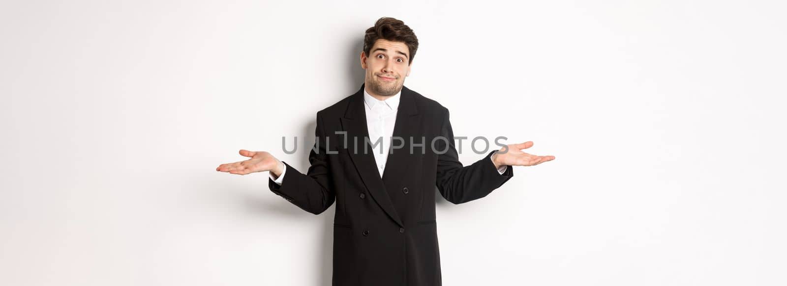 Image of handsome confused man in black suit, shrugging and looking clueless, dont know anything, standing over white background.