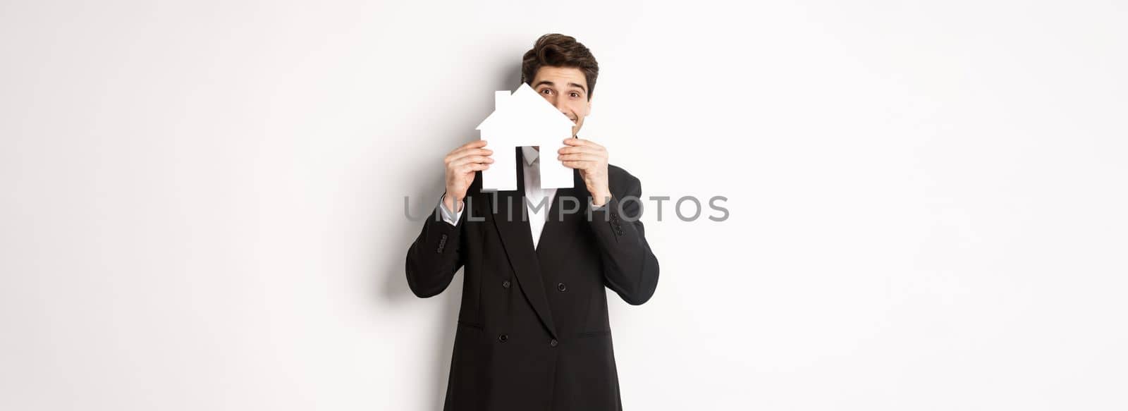 Image of handsome broker in black suit, showing house maket and smiling, selling homes, standing against white background.