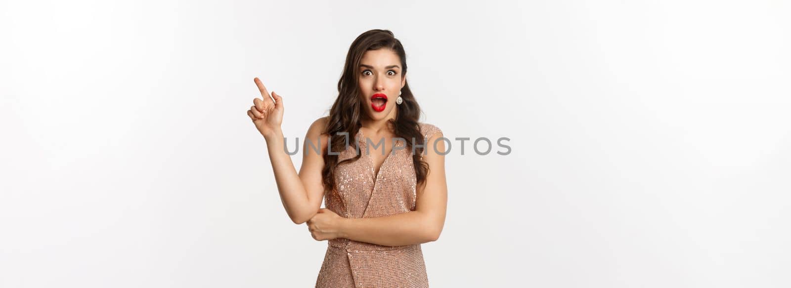 Surprised beautiful woman in luxurious dress and red lips, pointing finger right at promo and starring amazed at camera, standing over white background by Benzoix