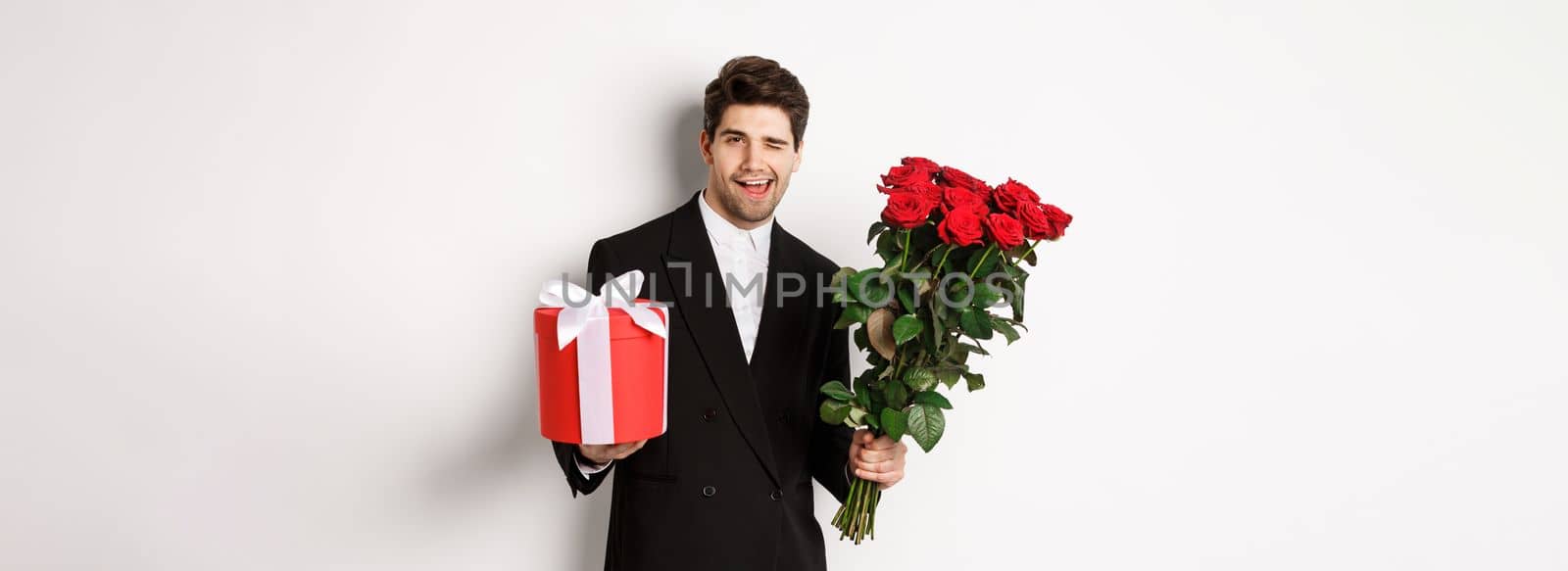 Concept of holidays, relationship and celebration. Handsome and confident man in black suit, going on a date, holding bouquet of roses and present, standing against white background.