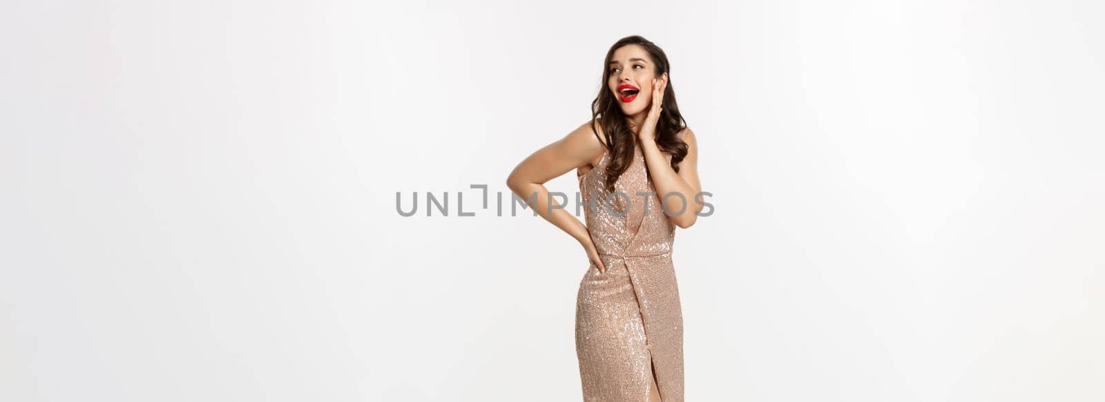 Party and celebration concept. Full-length of elegant woman in evening dress, standing near Christmas presents and looking surprised, standing over white background.