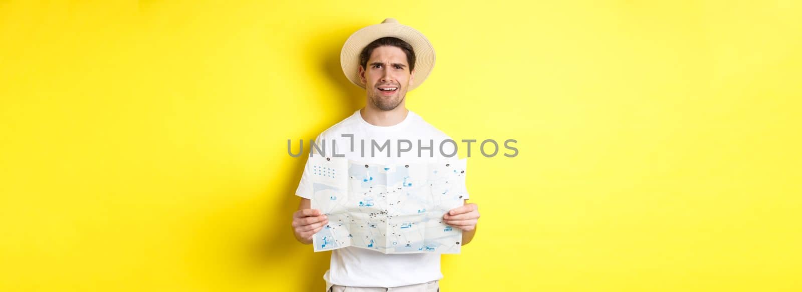 Travelling, vacation and tourism concept. Puzzled guy tourist cant understand map, looking confused at camera, standing against yellow background.