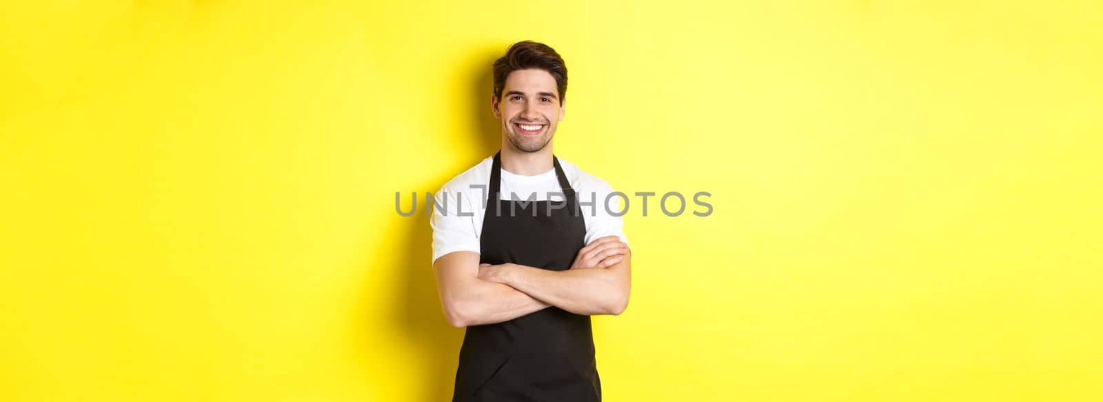 Smiling male waiter in black apron standing confident, cross arms on chest against yellow background.
