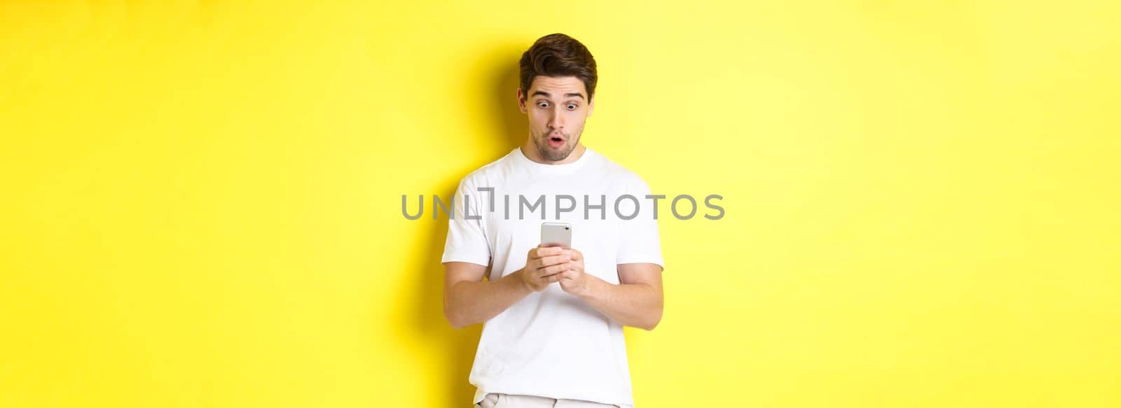 Man looking surprised in smartphone, reading message on cell phone, standing in white outfit against yellow background. Copy space