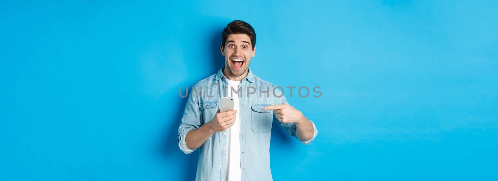 Concept of online shopping, applications and technology. Happy smiling man pointing finger at phone, standing amazed against blue background by Benzoix