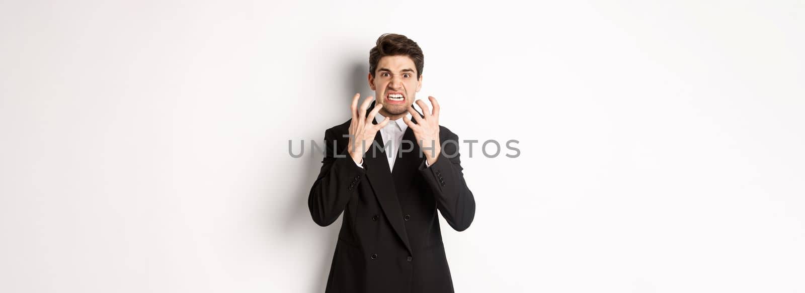 Image of angry businessman in suit, looking with furious expression and clenching fists, express hatred, standing mad over white background by Benzoix