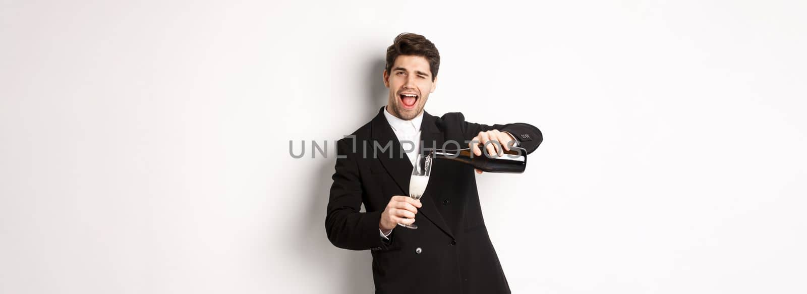 Handsome man in trendy suit pouring glass of champagne, celebrating christmas, smiling amazed and having fun, standing against white background.