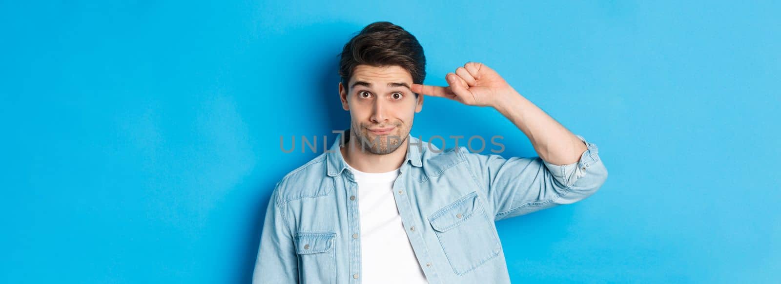 Close-up of man scolding for acting stupid or crazy, rolling finger on head and looking at camera, standing over blue background.