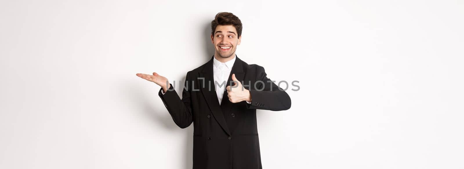 Handsome businessman in black suit, showing thumb-up and holding your product in hand over white copy space, standing against white background by Benzoix