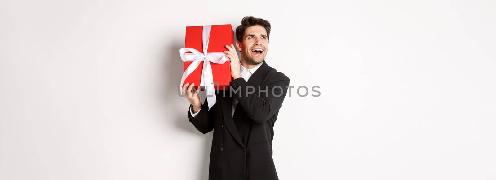Concept of christmas holidays, celebration and lifestyle. Image of excited man enjoying new year, shaking gift box to guess what inside, standing against white background.