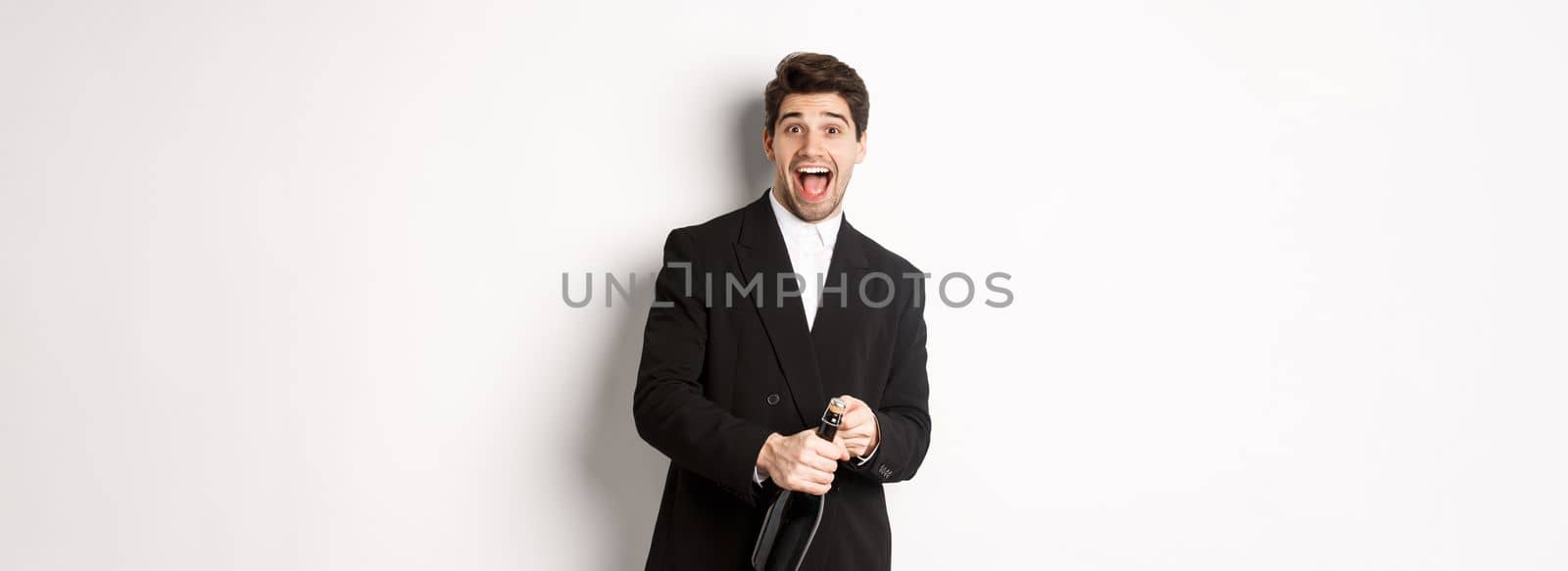 Image of attractive man in black suit having a party, celebrating new year and opening bottle of champagne, standing happy against white background by Benzoix