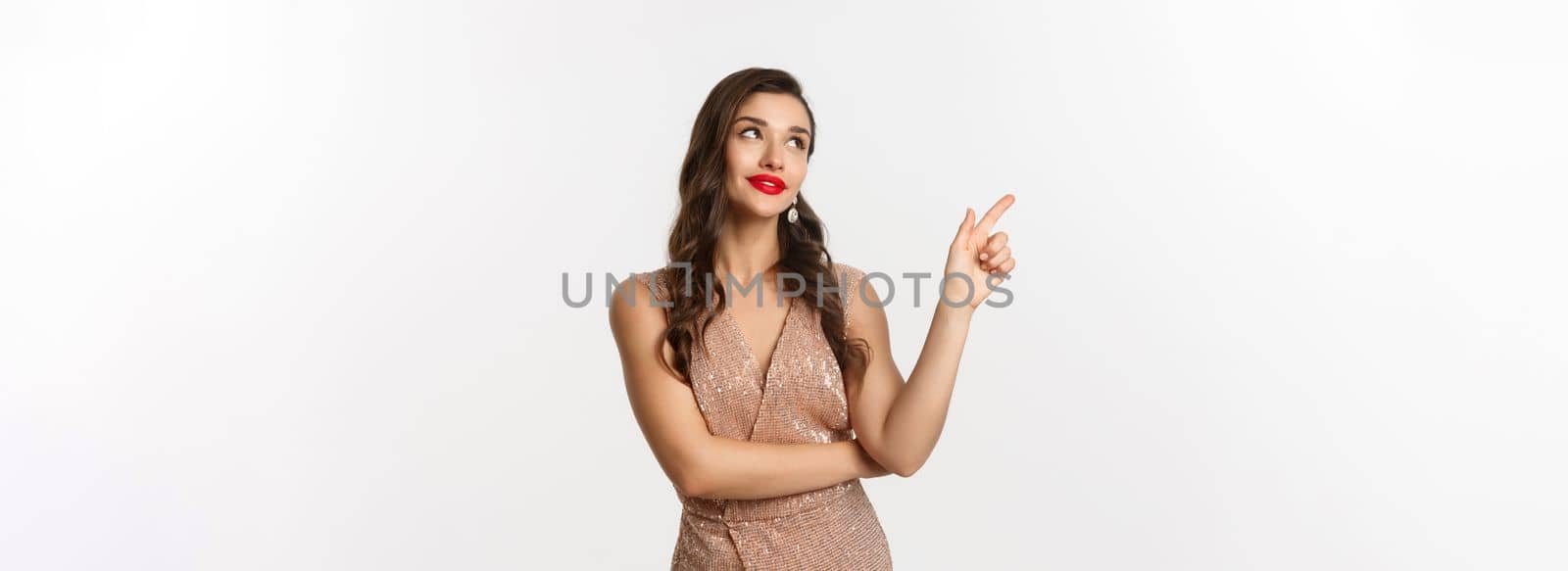 Christmas, holidays and celebration concept. Dreamy beautiful woman in party dress, pointing and looking left, thinking about new year party, white background.