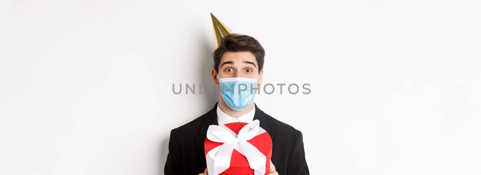Concept of party during covid-19. Close-up of happy guy in trendy suit and medical mask, celebrating christmas, holding present and smiling, standing over white background by Benzoix