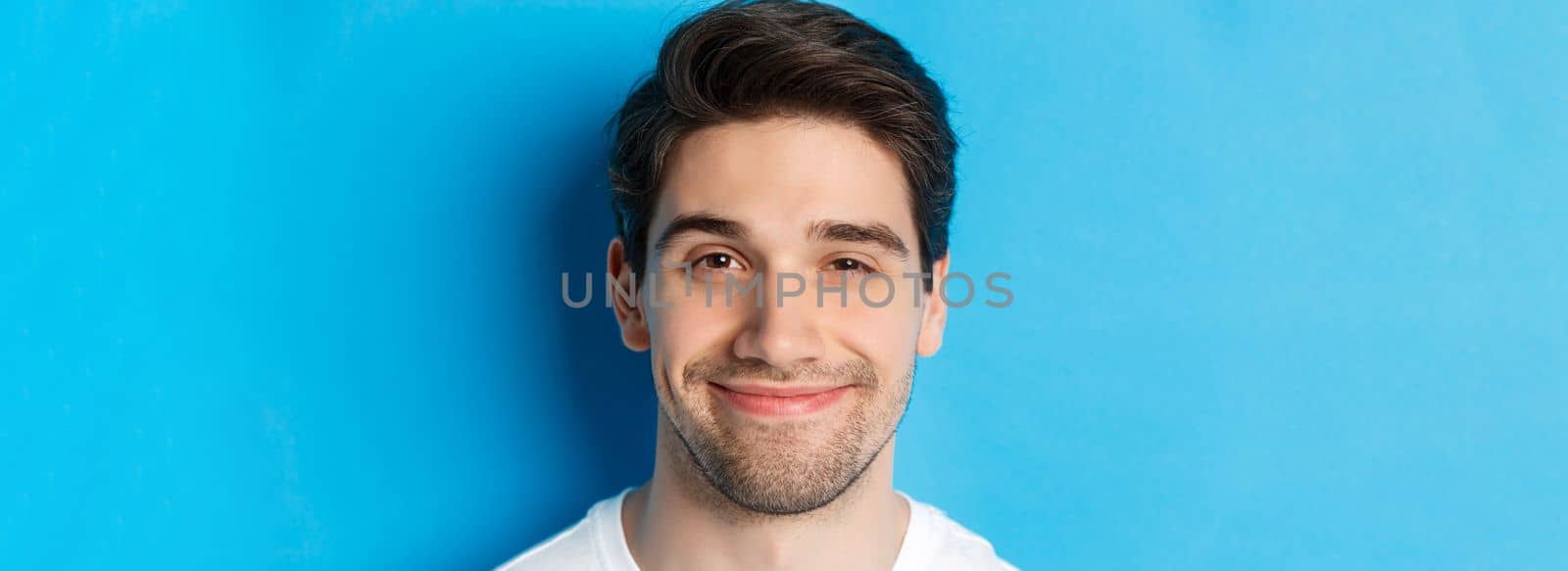 Headshot of attractive man smiling pleased, looking intrigued, standing over blue background.