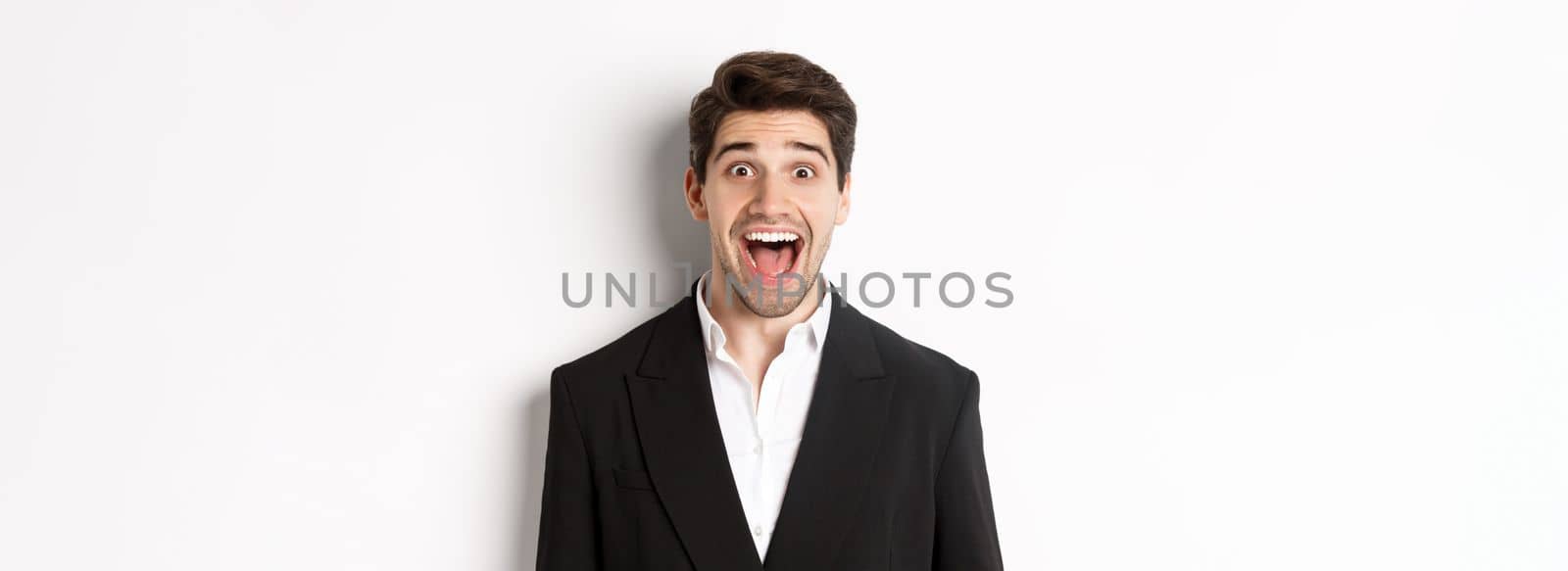 Close-up of attractive man in black suit, smiling amazed and looking at advertisement, standing over white background.