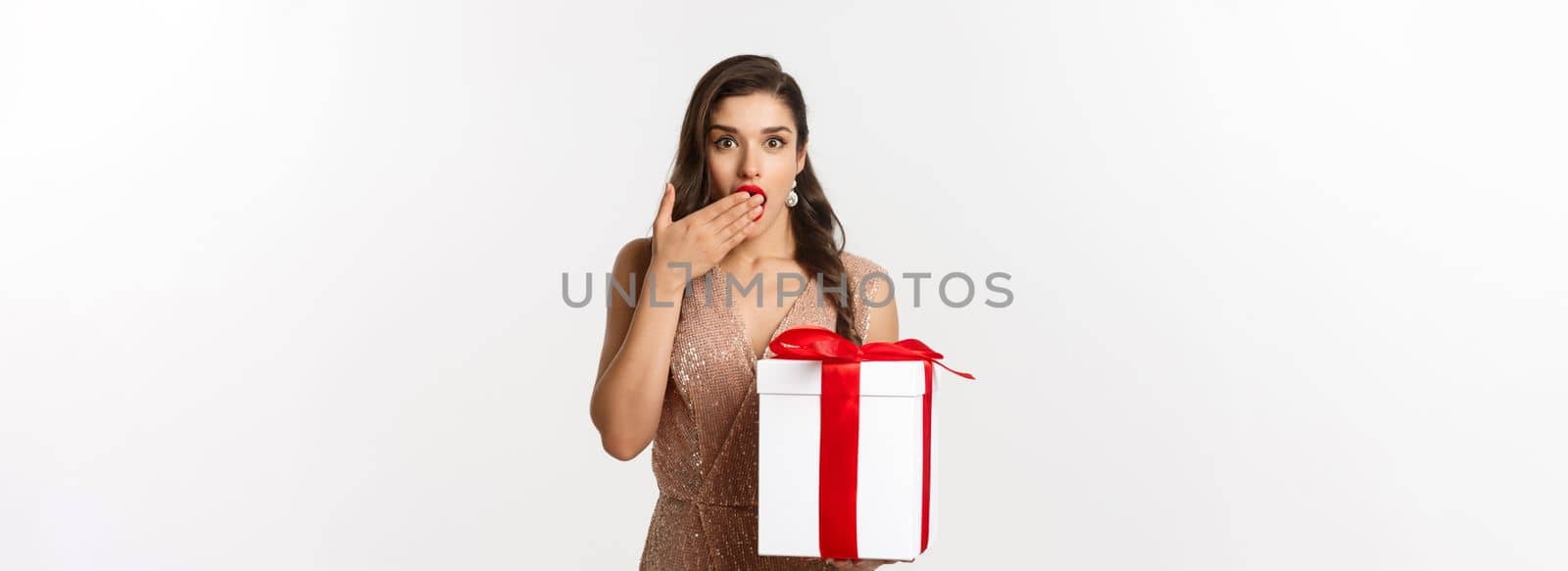 Merry Christmas. Beautiful woman looking surprised and holding gift, receiving new year presents, standing in luxury dress, standing over white background by Benzoix