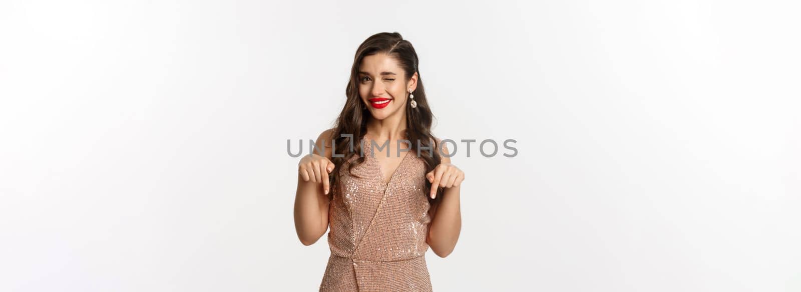 Elegant female model with red lips and earrings, pointing fingers down at christmas offer, winking and smiling, showing promo, standing in party dress over white background.