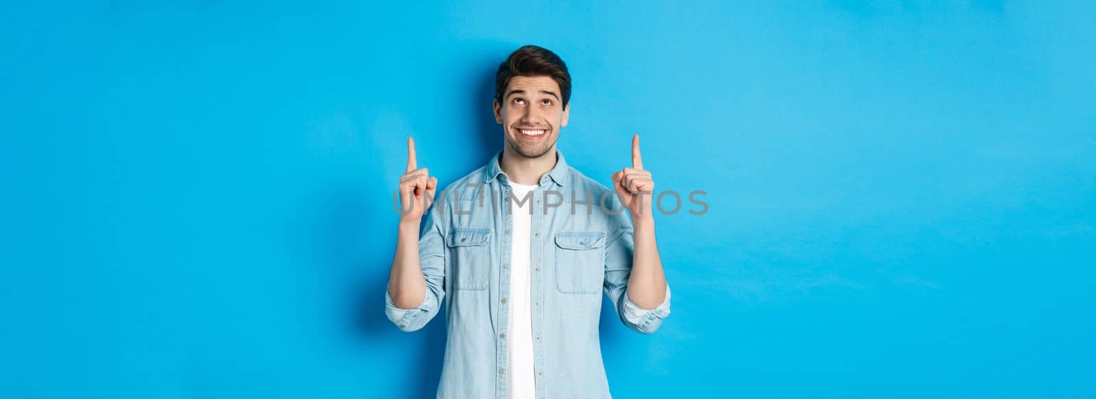 Portrait of handsome man in casual outfit, looking and pointing fingers up, showing banner on blue background.