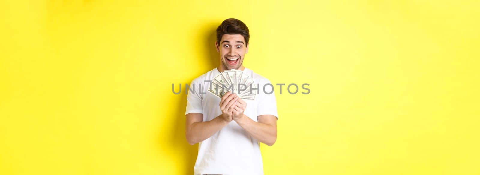 Happy man looking at money and smiling excited, winning prize, got bank loan, standing over yellow background.
