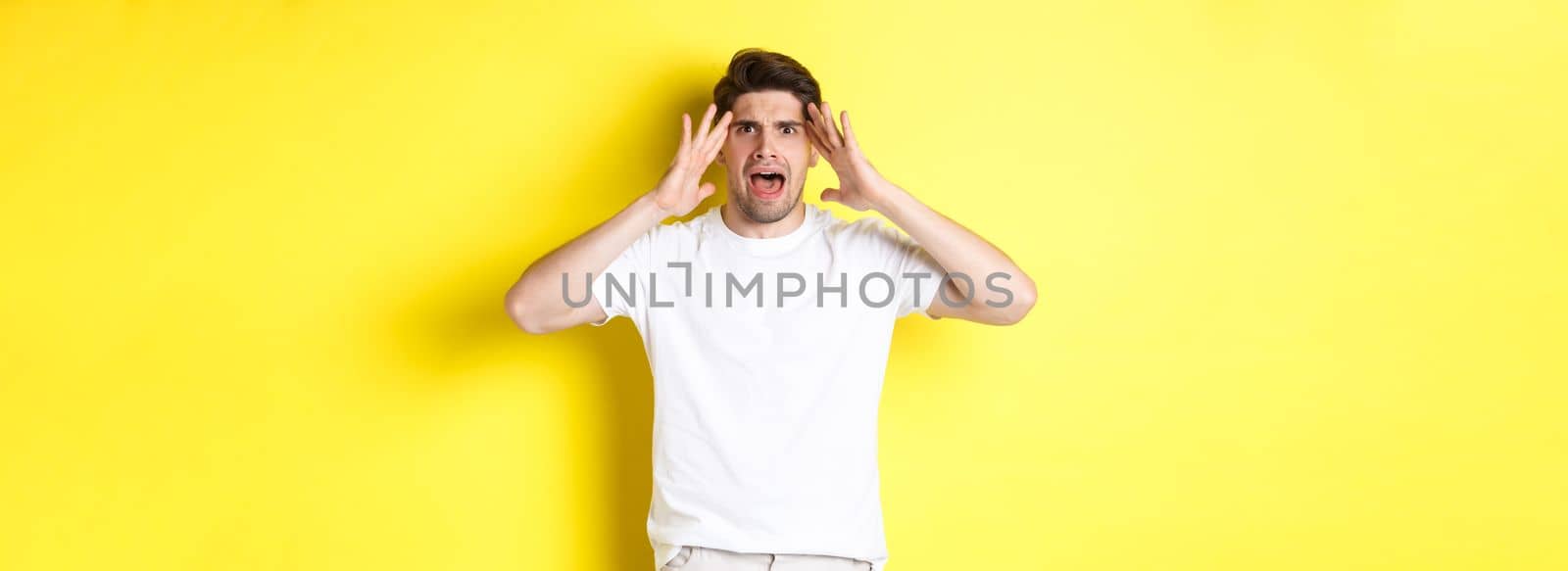 Frustrated guy looking alarmed, holding hands on head and feeling confused, panicking, standing over yellow background.