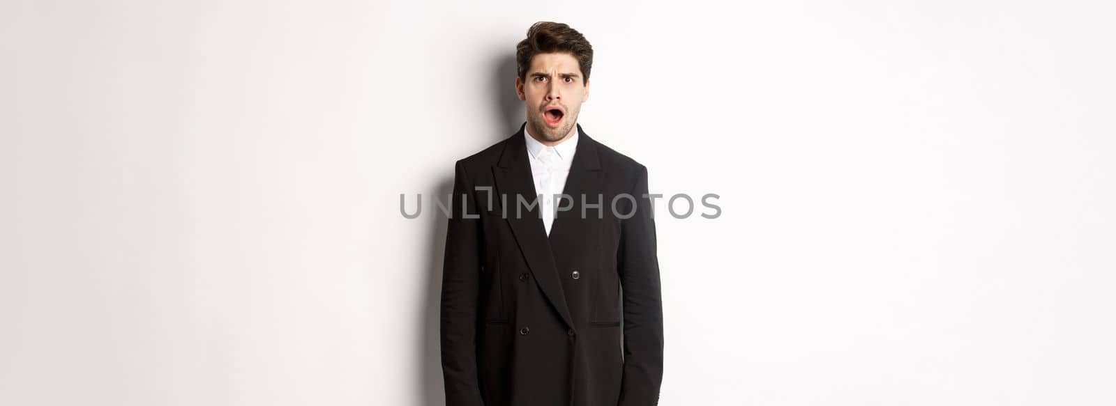 Portrait of shocked and startled handsome man in suit, drop jaw and looking in awe at camera, standing over white background by Benzoix