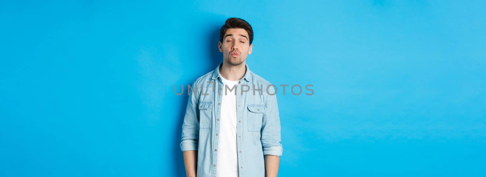 Handsome man waiting for kiss, pucker lips and close eyes while standing against blue background.
