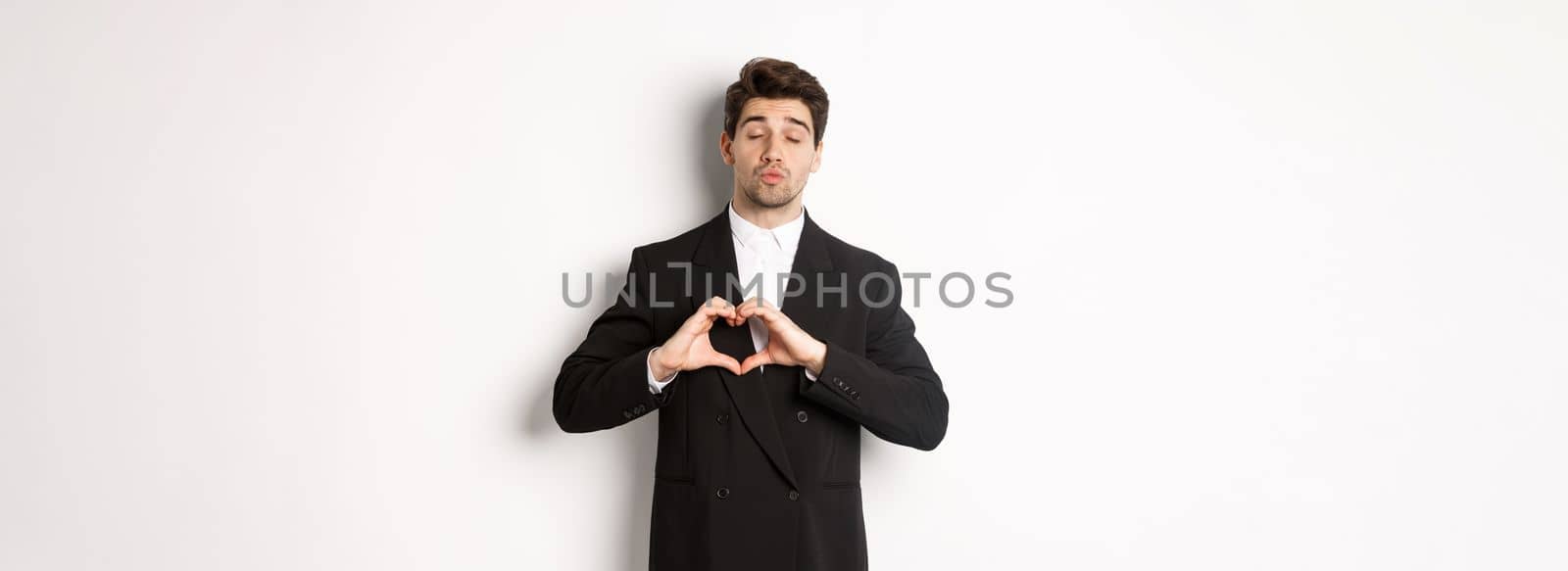 Image of handsome groom in black suit, showing heart sign, close eyes and pucker lips, waiting for kiss, standing against white background by Benzoix